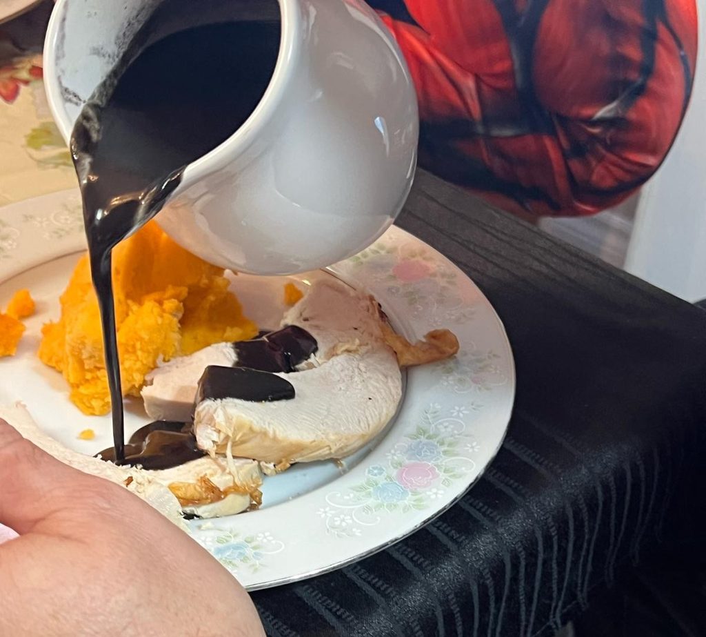 Black gravy being poured from a white gravy boat onto sliced turkey beside orange side dishes.
