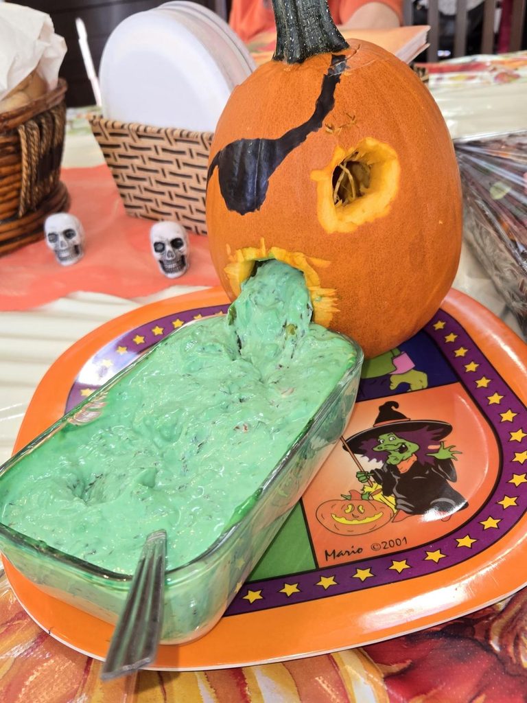 Pumpkin with a painted eye patch and green pistachio pudding salad spilling from its mouth into a serving dish.